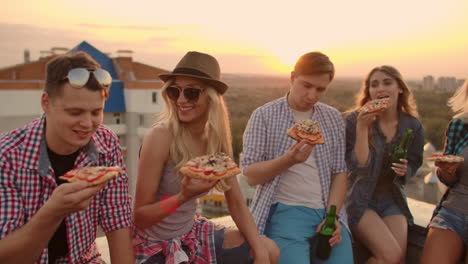 Young-people-at-sunset-on-the-roof-enjoy-pizza-and-beer.-They-sit-and-talk-to-each-other.